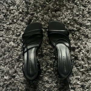 Svarta klackar från Pull&Bear - Stilrena svarta klackar från Pull&Bear med tunna remmar och fyrkantig tå. Skorna har låg klack och platt sula, perfekt för en trendig look. Tillverkade i syntetmaterial med minimalistisk design som passar till många outfits.