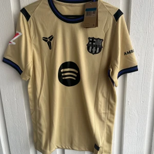 FC Barcelona Lamine Yamal bortatröja - Snygg FC Barcelona bortatröja i beige med svarta detaljer och tryck. Kortärmad modell med blå och svarta ränder vid ärmslut och krage. Tryck med Lamine Yamal och nummer 10 på ryggen. Materialet är lätt och andas, perfekt för fotboll eller att bära till vardags.