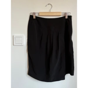 Svart silkeskjol från Les Petites - Svart kortkjol i 100% silke från Les Petites. Kjolen har en snygg dragkedja i sidan och diskreta veck framtill som ger en cool och stilren look. Perfekt för dig som gillar minimalistisk stil och vill ha något som känns lyxigt och bekvämt.