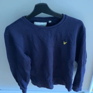 Mörkblå sweatshirt från Lyle & Scott - Säljer en mörkblå sweatshirt från Lyle & Scott med klassisk rund hals och lång ärm. Tröjan har en gul broderad fågel på bröstet och är gjord i mjuk bomull. Perfekt för chill dagar och enkel att matcha med jeans eller joggers.