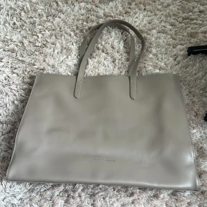 Beige axelväska från Smith & Canova - Stilren beige axelväska i slätt skinn från Smith & Canova. Väskan har dubbla handtag, silverfärgad dragkedja på baksidan och lila fodrad insida med flera fack. Perfekt för dig som vill ha plats för allt och ändå se chic ut.