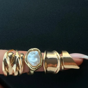 Set med chunky guldiga ringar - Fyra statementringar i guldton, alla med unik organisk form. En ring har en vit pärlliknande detalj i mitten, de andra har vågiga och skulpturala former. Perfekt för dig som gillar trendiga accessoarer med lite edge.