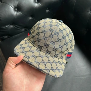 Gucci keps med GG-mönster och röd-blå rand - Exklusiv keps från Gucci i beige med mörkblått GG-monogram över hela ytan. Kepsen har en böjd skärm och ikoniska röd-blå ränder på sidan. Tillverkad i bomullsmix med ljus insida och klassisk passform. Perfekt för dig som vill sticka ut med lyxig streetstyle.