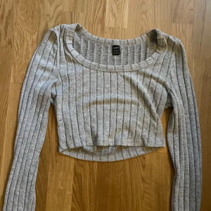 Grå ribbad croppad topp från SHEIN - Grå långärmad croppad topp från SHEIN med bred, rundad urringning. Toppen är ribbad med vertikala ränder och har en slim passform som sitter snyggt på kroppen. Perfekt att styla med höga jeans eller kjol för en chill look.
