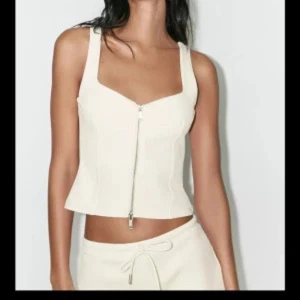 Vit korsett-topp med dragkedja - Snygg vit korsett-topp med croppad passform och breda axelband. Toppen har en markerad v-ringning och en silverfärgad dragkedja framtill som ger en cool vibe. Tillverkad i ett mjukt bomullsmaterial som känns skönt mot huden. Perfekt att styla med jeans eller kjol.