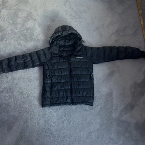 Svart dunjacka med huva från Peak Performance - Peak Performance dunjacka i mörktblå med huva och dragkedja framtill. Jackan har quiltad design, två sidofickor med dragkedja och justerbar huva. Perfekt för kalla dagar och har en clean, sportig look som funkar till streetwear. Materialet är syntet och dun.