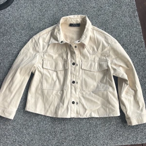 Beige skinnjacka från Bershka M - Snygg beige skinnjacka från Bershka i storlek M. Jackan har två stora bröstfickor med lock, svarta tryckknappar och klassisk krage. Materialet är mjukt och jackan har en rak, lite boxig passform. Perfekt för dig som vill ha en clean och stilren look.