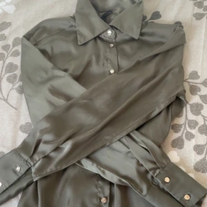 Olivgrön skjorta i silk  - Snygg olivgrön skjorta med silkeslen känsla och klassisk krage. Skjortan har långa ärmar med knappar vid manschetterna och knäppning framtill. Bara använts en gång då den är inte direkt min still längre.