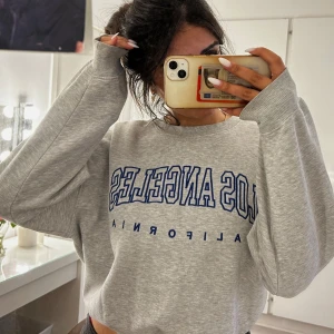 Grå sweatshirt Los Angeles BikBok - Grå sweatshirt från BikBok med blått broderat Los Angeles-tryck på bröstet. Oversized passform, rund halsringning och långa ärmar. Mjuk insida i fleece och ribbade muddar vid ärmslut och nederkant. Perfekt för en chill och sportig look.