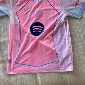 Barça Lamine Yamal #10 rosa Nike tröja - Säljer en FC Barcelona fotbollströja med Lamine Yamal och nummer 10 på ryggen. Tröjan är rosa med ljusblå detaljer, har korta ärmar och är tillverkad i ventilerande polyester. Nike-logga och Barça-emblem i holografisk stil, samt Spotify och UNHCR tryck. Perfekt för dig som älskar fotboll och stil.