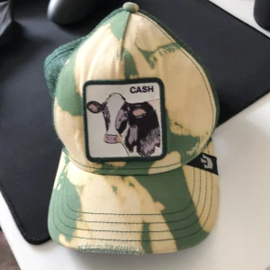 Goorin Bros keps med ko-tryck - Snygg keps från Goorin Bros 'The Farm' med grönt och beige camouflage-mönster. Framtill finns ett patch med en ko och texten 'CASH'. Kepsen har mesh baktill och justerbar snapback. Perfekt för dig som vill sticka ut med en unik accessoar.