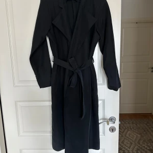 Svart trenchcoat från American Apparel - Svart trenchcoat från American Apparel med klassisk krage och bälte i midjan. Lång modell med raka ärmar. 