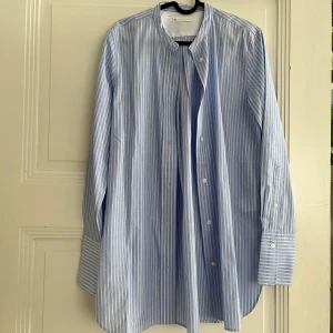 Blåvit randig skjorta från Arket - Snygg blå och vit randig skjorta från Arket i bomullspoplin. Skjortan har klassisk rak passform, ståkrage och långa ärmar med breda manschetter. Perfekt för en clean och stilren look. Knappar framtill och diskret logga i nacken.