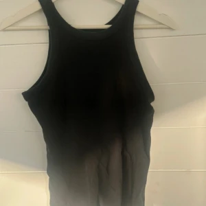 Svart ribbad linnetopp från 157 - Svart ribbad linnetopp från 157 i 100% bomull. Toppen har en tight passform med högt skuren halsringning och ärmlös design. Perfekt att styla med jeans eller kjol för en clean look.