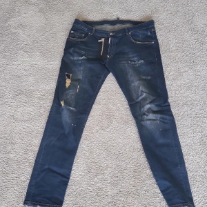 Dsquared2 blå skinny jeans med dragkedja - Snygga blå jeans från Dsquared2 med slitna detaljer och coola färgstänk. Modellen är skinny fit med en stor, synlig dragkedja framtill och klassiska fem fickor. Jeansen har en mörk tvätt och är tillverkade i jeansmaterial.