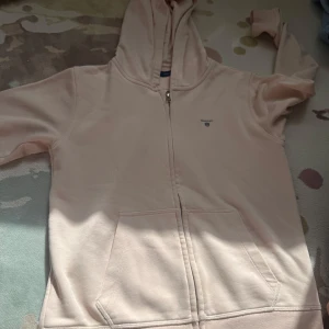 Ljusrosa hoodie från GANT med dragkedja - Säljer en ljusrosa hoodie från GANT med dragkedja framtill och huva. Tröjan har två stora fickor fram och diskret GANT-logga på bröstet. Tillverkad i mjuk bomull, perfekt för chill dagar. Klassisk och enkel design som funkar till det mesta.
