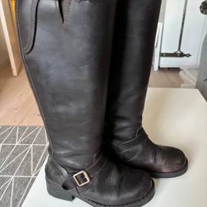 Bruna höga boots från Johnny Bulls - Snygga mörkbruna höga boots från Johnny Bulls i äkta skinn. Klassisk modell med rund tå, låg klack och spänne i metall vid ankeln. Grov sula för extra grepp och stilren look, perfekta till jeans eller kjol. Säljer nu pga att de bara stått i garderoben i flera år, köparen står för frakten.