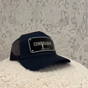 Mörkblå keps John Hatter & Co - Snygg mörkblå keps från John Hatter & Co med svart och silverfärgad metallskylt framtill där det står 'CONSIGLIERE'. Kepsen har mesh på sidorna och justerbar snapback baktill. Perfekt för dig som vill sticka ut med en cool accessoar. Aldrig använd!       Vid frågor eller funderingar är det bara att höra av sig! Säljes även på Vinted 