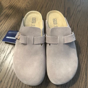 Birkenstock sandaler - Snygga sandaler från Birkenstock i beige mocka med klassisk slip-in design och justerbart spänne. Skorna har en mjuk innersula för extra komfort och en stilren look som passar perfekt till avslappnade outfits.