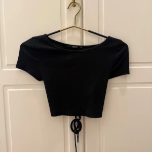 Svart topp - Svart crop top med snörning i ryggen från Bik Bok. Kortärmad modell med rund halsringning. Storlek XS. 