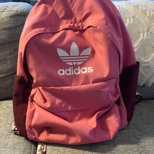 Rosa och vinröd Adidas ryggsäck - Säljer en snygg ryggsäck från Adidas i rosa och vinrött med stort vitt logotryck framtill. Väskan har ett stort huvudfack, framficka med dragkedja och vadderade axelband med Adidas-tryck. Perfekt för skolan eller träningen!