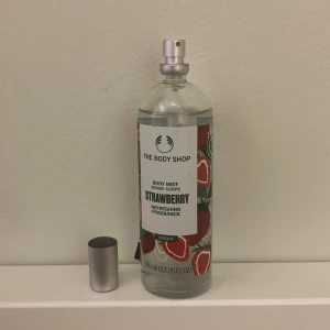 The Body Shop Strawberry Body Mist - Fräsch body mist med jordgubbsdoft från The Body Shop. Flaskan är genomskinlig i glas med ett färgglatt etikettmotiv av röda jordgubbar och gröna blad. Silverfärgad spraykork. Vegansk och innehåller 100 ml. Perfekt för dig som gillar söta och fruktiga dofter.
