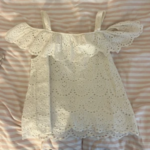 Vit spetstopp Holly & Whyte - Superfin vit topp från Holly & Whyte med broderad spets och volang upptill. Toppen har axelband och offshoulder-detalj, perfekt för sommarens varma dagar. Söt och luftig modell med vackert mönster och scallop-kant nedtill.