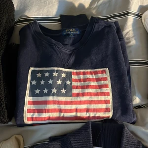  Polo Ralph Lauren - Mörkblå sweatshirt från Polo Ralph Lauren med stor amerikansk flagga framtill. Klassisk rund halsringning och mjukt material som känns skönt mot huden. Perfekt för dig som gillar stilrena och ikoniska plagg med en sportig touch. Size 14-16 år passar s man