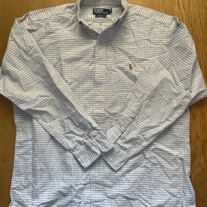 Rutig skjorta från Polo Ralph Lauren - Klassisk rutig skjorta från Polo Ralph Lauren i vitt och blått. Skjortan har lång ärm, knappar framtill och ikonisk broderad logga på bröstet. Tillverkad i 100% bomull för en skön och luftig känsla. Perfekt till jeans eller chinos.