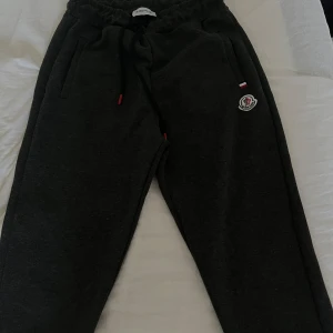 Svarta mjukisbyxor från Moncler - Mörkgrå mjukisbyxor från Moncler med dragsko i midjan och broderad logga på benet. Byxorna har sidofickor och en relaxed passform. Snygga detaljer med svarta snören och små röda accenter. Använt 2 gånger bara 