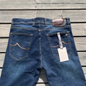 Jacob Cohën blå jeans med läderpatch - Snygga mörkblå jeans från Jacob Cohën med klassisk femficksdesign, kontrastsömmar och läderpatch bak i midjan. Jeansen har raka ben och är tillverkade i mjukt bomullsjeansmaterial. Detaljer som broderad logga och dekorband vid bakfickan.