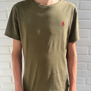 Olivgrön t-shirt från Ralph Lauren - Snygg olivgrön t-shirt från Ralph Lauren med klassisk röd broderad logga på bröstet. T-shirten har rund halsringning och korta ärmar. Tillverkad i mjuk bomull som känns skön mot huden. Perfekt för en clean och avslappnad stil.