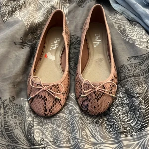 Rosa ballerinaskor med ormmönster från Duffy - Snygga ballerinaskor från Duffy i ljusrosa med ormmönstrad yta och dekorativ rosett framtill. Skorna har rund tå och platt sula, vilket gör dem både stilrena och bekväma. Perfekta för dig som gillar detaljer och vill sticka ut med ett coolt mönster.