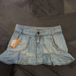 Ljusblå lågmidjad jeanskjol med volang - Supersöt ljusblå jeanskjol med volangkant och slitna detaljer. Kjolen har klassiska fickor fram och bak, bälteshällor och knappstängning. Perfekt för dig som gillar en chill och trendig look med lite edge.