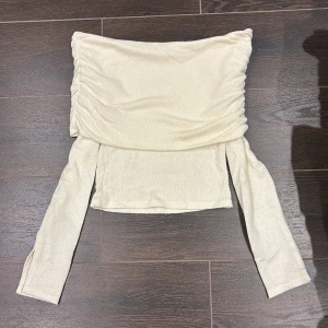 Offshoulder topp i beige från Gina Tricot - Trendig offshoulder topp från Gina Tricot i ljusbeige stickat material. Långa ärmar och snygg rynkdetalj längs axlarna ger en cool och modern look. Perfekt att styla med höga jeans eller kjol för en chic vibe.