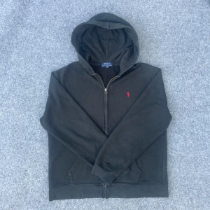 Svart hoodie från Polo Ralph Lauren - Svart hoodie från Polo Ralph Lauren med dragkedja och huva. Klassisk röd logobrodering på bröstet och två fickor framtill. Tillverkad i mjuk bomull, perfekt för chill dagar. Snygg och enkel design som funkar till det mesta.