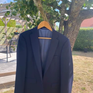 Mörkblå kavaj Dressmann slim fit - Snygg mörkblå kavaj från Dressmann i slim fit-modell, storlek 46. Klassisk design med fyra svarta knappar vid ärmslut, diskreta fickor och stilren insida med röd kant. Perfekt för dig som vill ha en stilren look.