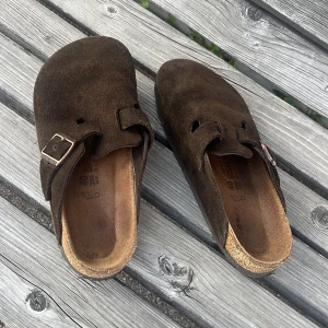 Birkenstock Boston mocka brun 40 - Säljer ett par Birkenstock Boston i brun mocka med klassisk spänndetalj på ovansidan. Skorna har en rund tå och platt sula, samt en mjuk innersula för extra komfort. Perfekta för dig som gillar chill och stilren look.