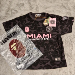 Inter Miami x BAPE - Snygg svart och grå camo fotbollströja från A Bathing Ape i samarbete med Inter Miami. Tröjan har korta ärmar, rosa MIAMI-tryck och broderad klubbmärke på bröstet. Tillverkad i mjuk bomull med klassisk BAPE-logga och detaljer i rosa. Den passar xs och s. Priset är ej hugget I sten