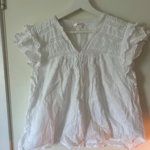 Vit blus med volang från Gina Tricot - Supersöt vit blus från Gina Tricot i 100% bomull. Blusen har v-ringning, volangdetaljer vid ärmarna och ett luftigt, flowy fall. Perfekt för dig som gillar romantisk och avslappnad stil. Passar till jeans eller kjol för en fräsch look.