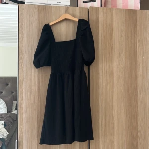 Svart klänning med puffärm från H&M - Säljer en svart klänning från H&M i storlek XS. Klänningen har puffiga korta ärmar, fyrkantig urringning och smockad överdel. Kjolen är lätt utställd och längden går till knäna. Är använd en gång