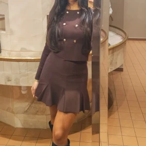 Mörkbrun kort kjol med volang med en snygg topp sett - Supersnygg mörkbrun kort kjol med långärmad överdel och dekorativa guldknappar framtill. Kjolen har volangkant som ger en extra touch. Passformen är normal och materialet känns mjukt och stretchigt, perfekt för dig som gillar stilrena detaljer. Nåde toppen och kjolen säljs tillsammans för ett bra pris 🤍