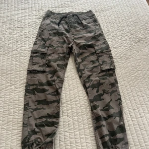 Cargobyxor med camouflage och snörning - Säljer ett par cargobyxor i camouflage-mönster med elastisk midja och snörning. Byxorna har stora fickor på sidorna och är perfekta för en avslappnad streetstil. Materialet känns som bomull och de har muddar vid bensluten. 