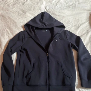 Mörkblå hoodie från Polo Ralph Lauren - Snygg mörkblå hoodie från Polo Ralph Lauren med dragkedja och huva. Tröjan har broderad logga på bröstet, praktiska fickor och snörning i huvan. Tillverkad i mjuk bomullsmix, perfekt för chill dagar eller när du vill ha en clean look. Nästan ny skick