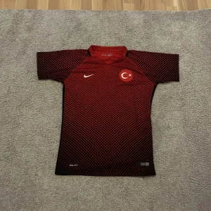 Turkiet Nike matchtröja röd-svart - Säljer en officiell Turkiet fotbollströja från Nike, modell 2016. Tröjan är röd med svarta rutor och har korta ärmar. Nike-logga och Turkiets emblem på bröstet. Tillverkad i Dri-FIT-material som andas och passar perfekt till träning eller match.