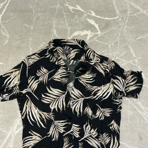 Svart mönstrad skjorta från H&M - Snygg svart skjorta från H&M med korta ärmar och beige tropiskt bladmönster. Skjortan har klassisk krage och knäppning framtill. Perfekt för dig som vill sticka ut med en cool och avslappnad stil.