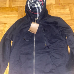 Svart hoodiejacka från Burberry - Svart hoodiejacka från Burberry med dragkedja och klassiskt rutig foder i huvan. Jackan har snörning i huvan, två fickor fram och är tillverkad i mjuk bomull. Perfekt för dig som gillar stilren design med en touch av lyx.