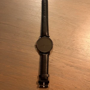 Svart minimalistisk klocka med läderband - Stilren svart klocka med rund urtavla och svart läderband. Urtavlan är helt svart utan siffror och har tunna visare för en clean look. Perfekt accessoar för dig som gillar enkel och modern design.