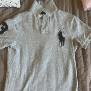 Grå pikétröja Polo Ralph Lauren S - Snygg grå pikétröja från Polo Ralph Lauren i slim fit. Klassisk krage, korta ärmar och knäppning framtill. Stor svart logga med polospelare på bröstet och svart siffra på ärmen. Perfekt för dig som gillar stilrena och sportiga plagg.