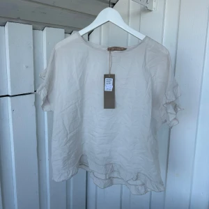 Beige blus med volangärm från Marta - Luftig och lätt beige blus från Marta med korta ärmar och volangdetaljer. Blusen har en avslappnad passform och rund halsringning, perfekt för en chill och stilren look. Materialet känns tunt och svalt, vilket gör den skön att bära under varmare dagar.